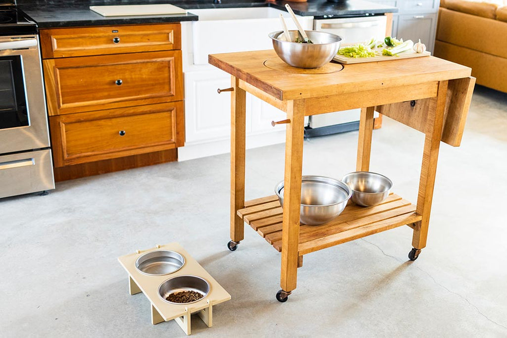 A kitchen area with a wooden food preparation cart. On the floor to the left of the cart is a Rise Pet Bowl Stand for large bowls, low rise height. On the bottom tier of the wooden food preparation cart are 3 Workhorse Stainless Steel Mixing Bowls. On top of the wooden food preparation cart is a single Workhorse Stainless Steel Mixing Bowl containing mixing utensils along with a single Workhorse HDPE cutting board with chopped vegetables on top.