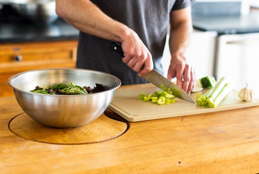 A Workhorse Stainless Steel Mixing bowl filled with salad to the left and a person chopping celery on a Workhorse HDPE Cutting Board on the right, both on a wooden food preparation surface.