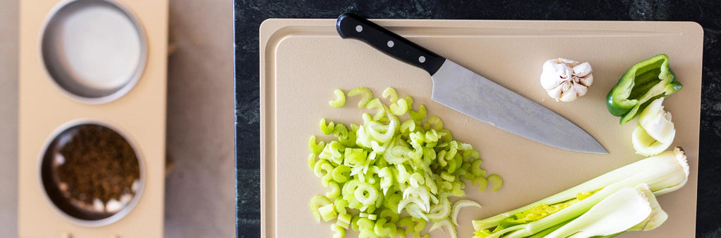 A view from above of a Workhorse HDPE cutting board with chopped vegetables and a chefs knife. Out of focus in the background is a Rise Pet Bowl Stand with 2 bowls - one containing kibble and the other water.