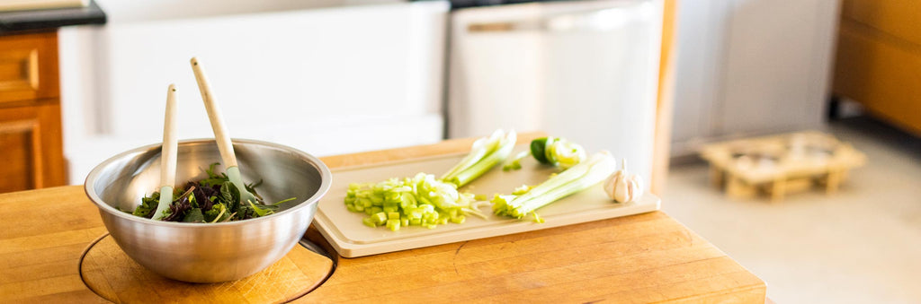 A Workhorse Stainless Steel Mixing bowl containing salad and mixing utensils rests on the left side of a wooden food prep surface with a Workhorse HDPE cutting board containing chopped vegetables resting on the right side of the wooden surface. Out of focus in the background is a kitchen scene with a Rise Pet Bowl Stand on the floor.