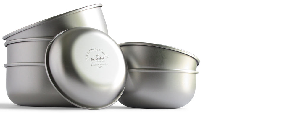 A stack of 3 large size stainless steel dog bowls and a stack of 2 medium size stainless steel dog bowls in the background. In the foreground is a single small size dog bowl in a vertical position with the bottom facing forward to show the Basis Pet laser mark and logo.