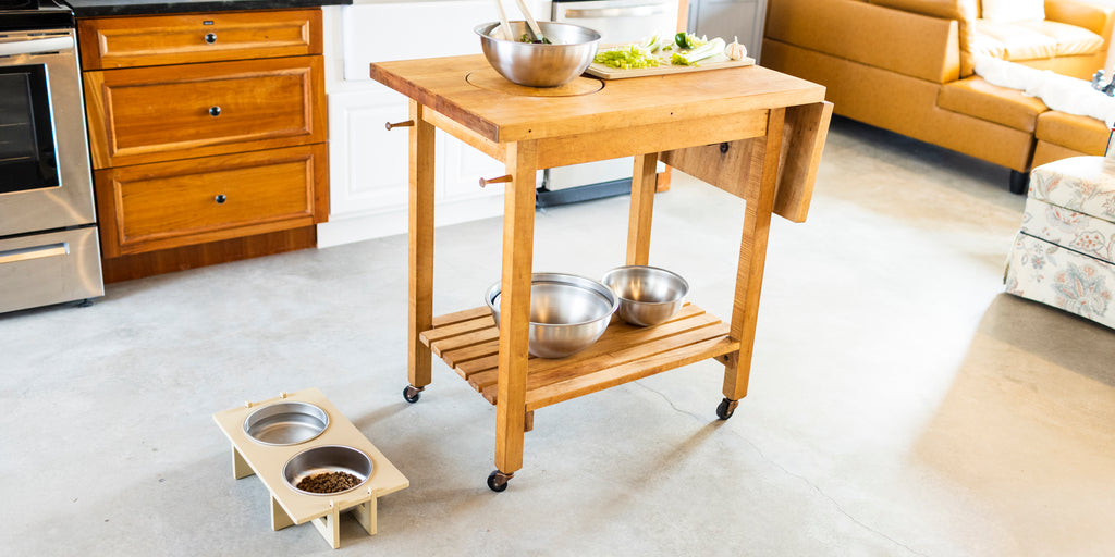 A wooden kitchen prep cart with a stainless steel bowl and cutting board on top. Variously sized stainless steel mixing bowls are stored on the cart shelf. A pet food stand with stainless steel bowls is located on the floor next to the cart station.