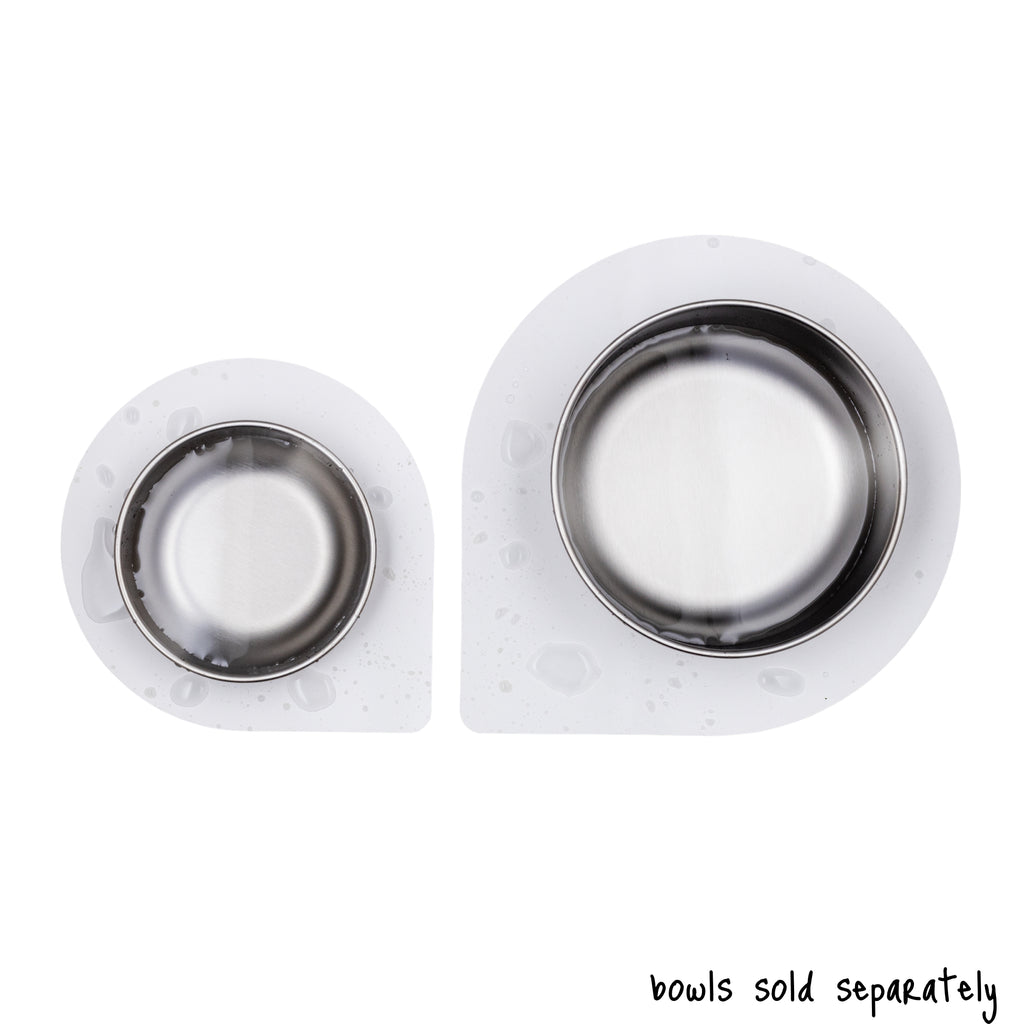 A small size Basis Pet stainless steel dog bowl on a regular size Ultra Grip mat on one side and a large size Basis Pet stainless steel pet bowl on a large size Ultra Grip mat on the other side. Both bowls contain water and are shown from above. Text reads "bowls sold separately".